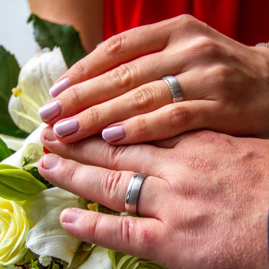 Hochzeitsplanung Hände mit Trauringen und Blumen, hinterlegt von einer roten Stoffoberfläche.